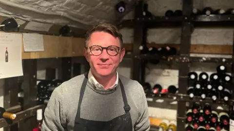 A bespectacled man wearing a grey jumper and apron smiles. In the background are shelves stacked with wine bottles.   