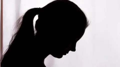 BBC A silhouette of a woman looking to the ground. She has a long ponytail and is stood against a white background. 