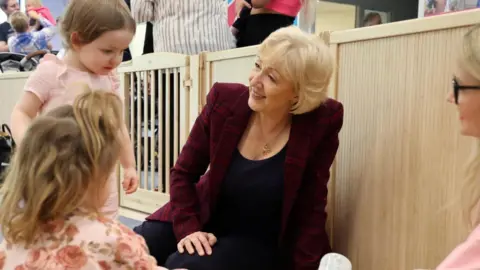 1001 Critical Days Foundation A person wearing a dark checkered jacket is seated on the floor inside a play area, interacting with young children. The setting includes a light wooden safety gate and a soft yellow mat. Several children are nearby, one holding a baby bottle, and another wearing a floral-patterned top. In the background, more people and children can be seen, suggesting a busy, child-friendly environment such as a nursery or playgroup.