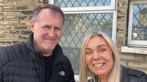 Grace Wood/BBC A man with short brown-grey hair wearing a black puffer jacket and a woman with long blonde hair wearing a black leather jacket smile at the camera.