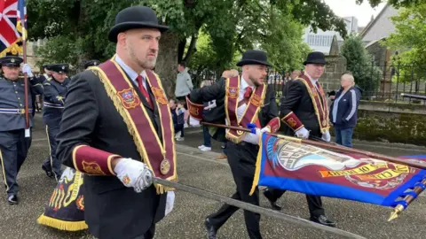 Three men wearing black suits and bowler hats walking in a parade. The man to the forefront of the photo is holding a sword and the man in the middle is holding a flag.