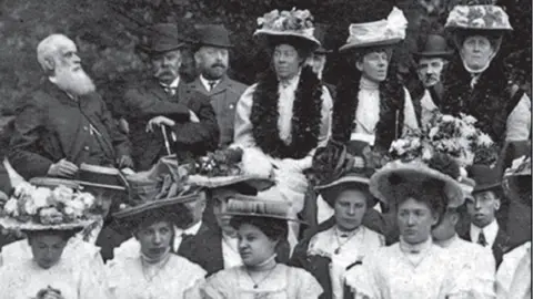 Thetford Town Council A black and white Edwardian photo of a group of men and women. Allan Minns is in the back row, second left, with folded arms, wearing a hat, holding a stick. He has a drooping large moustache covering his upper lip. Ophelia Minns is the first woman on the right on the back row. She is wearing a white dress, has a floral garland around her neck and is wearing a large flower-covered hat