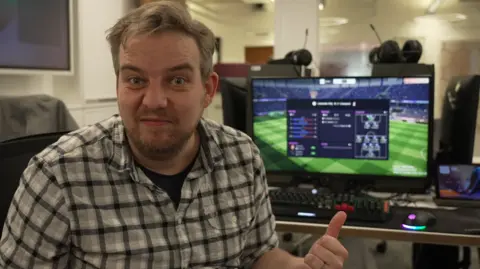 Tom Gerken sits at a computer. He is looking directly at the viewer and gesturing to the computer behind him, on which appears to be a video game screen.