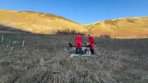 Scottish Water Two people in red jackets and trousers and black boots work on a drone with a backdrop of hills in southern Scotland