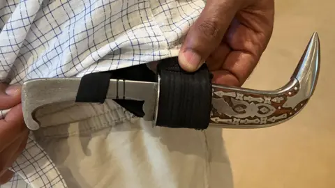 A Kirpan is held in a holster attached to a man's trousers. The blade is covered by a wooden sheath.