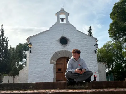 Alex Batty sitting cross‑legged on a low stone wall holding a mobile phone, with a plastic drinks bottle placed beside them. Behind, a small whitewashed church with a wooden arched door, a circular window with metal grille, and a bell housed in a simple open belfry topped by a cross. Two black wall-mounted lanterns flank the entrance. Trees and sky form the background.
