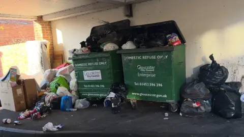 Two large green bins, which are overflowing with full black bin bags, carrier bags, and cardboard boxes. A large amount of waste has been deposited beside the bins, due to them being full. They are on a wide section of pathway beneath an archway under a block of flats.