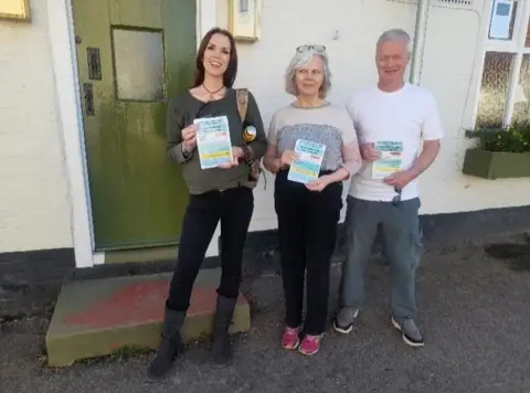 Communities Against Factory Farming Two women and a man stand outside a building with a green door and white walls, looking towards the camera, holding up flyers.