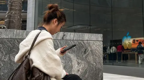 A woman uses a smartphone while sitting outside an Apple Store in Chongqing, China.