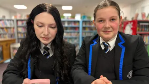 Two girls wearing school uniform with black blazers and blue edging standing together in a library with one holding a pink mobile