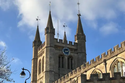 A church clock tower stands in front of blue skies with white wispy clouds. The time reads a quarter past two on a blue clock face. A black street lamp and a few tree branches can be seen in the bottom right corner of the photo.