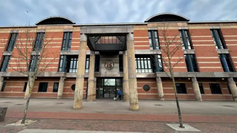 BBC Teesside Crown Court. A large three storey building made from red brick with long narrow black windows. Four large round yellow stone columns support a pyramid shaped glass roof above the front door.