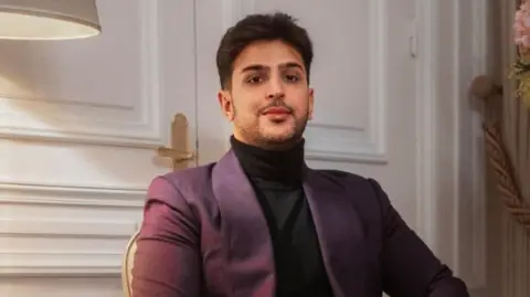 Varun Datta A man wearing a black turle neck jumper and a purple jacket. He is looking directly at the camera. 