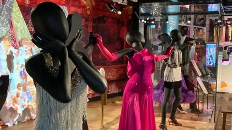 Black faceless mannequins lined up in poses wearing Strictly Come Dancing costumes with tassels and sparkles in silver and hot pink
