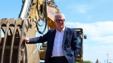 West Midlands mayor Richard Parker wears a white shirt and dark blue suit. He has white hair and black-rimmed glasses. He is leaning against the bucket of a yellow construction digger.