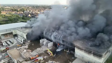 London Fire Brigade Smoke billowing out from a large warehouse with fire trucks and the brigade trying to put it out.
