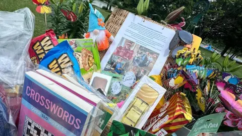 Rachel Davies A number of items are seen in close-up on a table. There are crossword books protected in clear plastic bags, snacks in wrappers, a sample of aftershave, garden seeds and butterfly ornaments