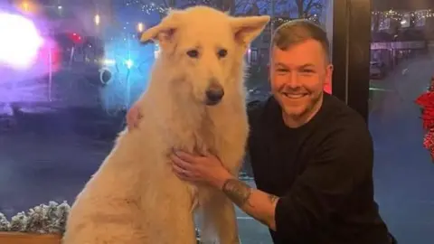 A young man with short hair and tattoos on his forearm stands with his arms around a large white dog. He is smiling. 