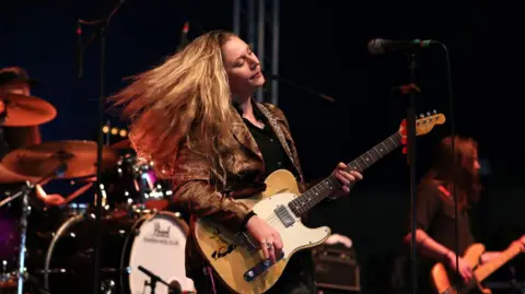 A woman playing a guitar, her hair is flying behind her and the stage is backlit with a red light. There are drums in the background of the image. 