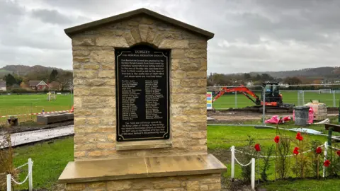 A war memorial with a small garden behind it which is lined with poppies and a white low chain fence. The memorial itself is made of cotswold stone and has a large black plaque on it with the names of 44 soldiers who died in World War One and World War Two. 