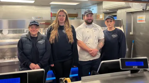 Three women and a man in branded uniform stand in a fish and chip shop. Behind them is the stainless steel range and in front of them are digital till displays.