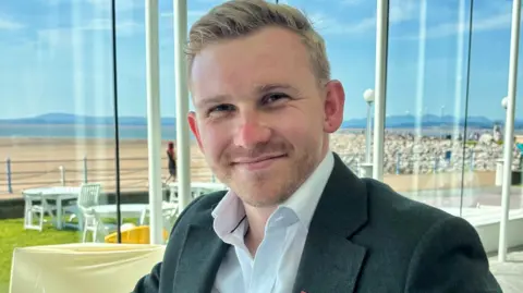Dewey Lord with short blond hair and stubble wearing a suit sits inside the Midland Hotel in Morecambe with a seaview of Morecambe Bay beach behind him through the windows. It is a hot day and the tide is out. Hills are seen in the background. He is smiling.