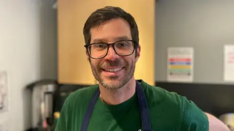 Rob Lewis wears a green T-shirt and a blue apron. He's also got glasses on and has brown hair. He's smiling at the camera and in the background is a kitchen. 