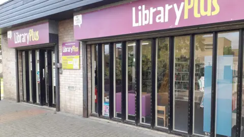The outside of the library, which has large floor to ceiling glass windows and a purple sign saying Library Plus