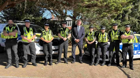 The PCC poses in the centre of a line up of officers in front of a police car and police 4x4.