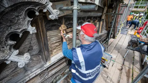 National Trust A man repairing some woodwork. He is wearing a red hat and a blue top and blue jacket with writing on it. 