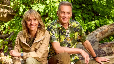 Michaela Strachan and Chris Packham are sitting outside on an old tree which is in a forest. They are smiling at the camera. 