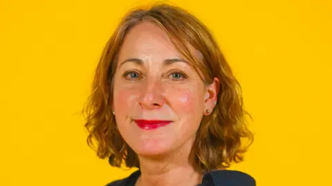 University of Suffolk A headshot image of Prof Emma Bond who is smiling at the camera. She has wavey red hair which is cut just above her shoulders. She stands in front of a bright yellow background. 