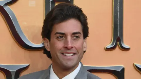 James Argent smiling at a red carpet event. He has dark hair swept backwards and is wearing a grey suit jacket and white shirt.