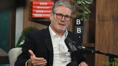 BBC  Sir Keir Starmer sitting behind a microphone at the BBC, gesturing with his hand, with various BBC logos on the wall behind him