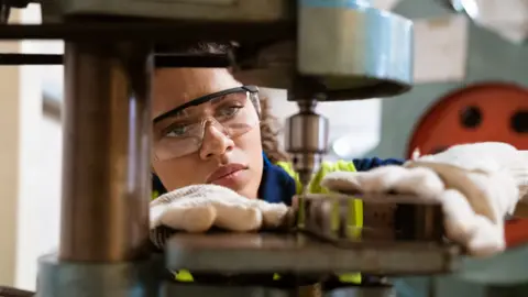 Getty Images  Young female apprentice wearing protective goggles and gloves works on a machine 