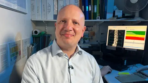 Professor Alex Comninos in a light-coloured shirt sits at a desk in an office, facing the camera. Behind him, shelves are filled with binders and documents, and a computer monitor displays a detailed spine image alongside a coloured graph. A desk surface holds items including a highlighter and notepad, while office equipment such as a fan and stacked files appear in the background.