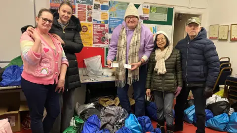 BBC Kerry Warwick wearing a bright pink cardigan and glasses stood with volunteers inside King Edward Park pavilion