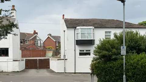 Fence on street leads into a car park with broken fencing behind it - white buildings line the street