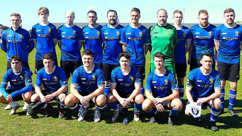 Robert Kirkland Alderney FC players pose for a team photo before playing a match on a sunny day. They are wearing their blue home kit. The goalkeeper is wearing a green kit.