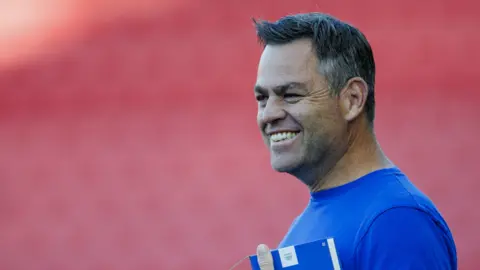 Bath director of rugby Johann van Graan smiling before a Prem Rugby mach, wearing a blue t shirt and holding a blue notebook.