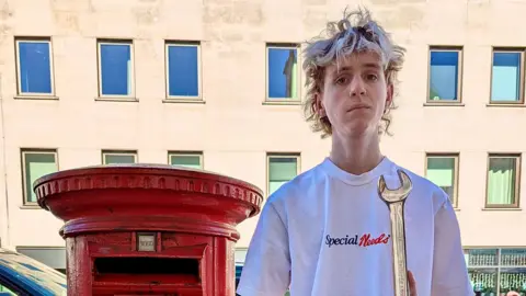 Ed Gold/Positive Futures Ethan holding a large metal spanner while standing next to a red pillar-style postbox. He has wavy blond hair and is in front of a tall white building.