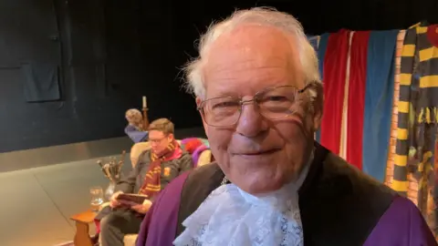 Luke Deal/BBC An elderly man with thin white hair wearing glasses and a purple and black outfit smiling into the camera. 