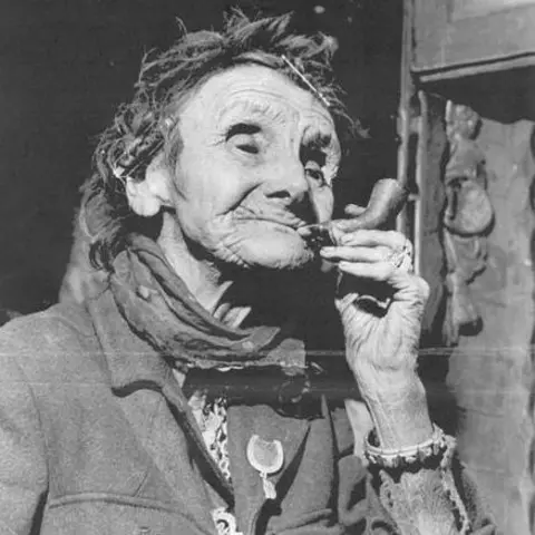 Walsall Archives A black and white image of Louisa Price. She is smoking a clay pipe and has a scarf around her neck. On her jacket is a lucky horseshoe and she has a heavy bracelet around her wrist