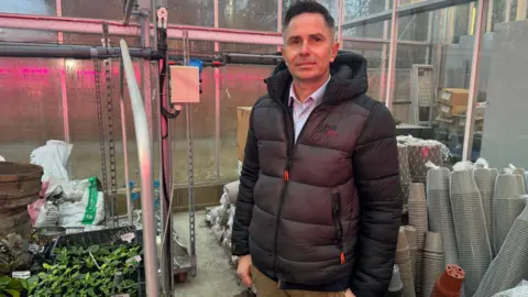 Alan Harvey, one of the Hadlow College lecturers stands near a series of trays holding bedding plants. He's wearing a grey coat and tan chinos.