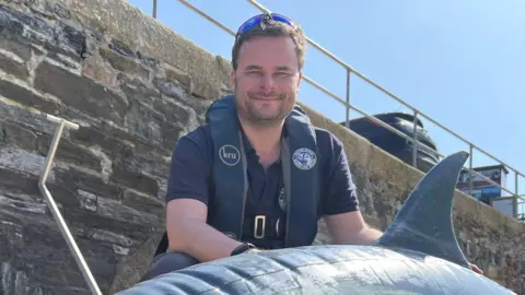 BBC A man is smiling towards camera he's wearing a blue short sleeved T.shirt. He's kneeling behind a blow up dolphin. The dolphin is closer to the camera. It's grey with a cream flank. It's resting on sand. A large harbour wall is in the background and you can see blue sky in the right top corner of the photo.