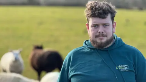 A man standing looking at the camera with a neutral expression. He is wearing a blue hoodie. He is standing in a field with some sheep behind him.