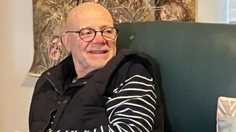 Malcolm wears a stripy navy and white top under a black gilet and sits on a green sofa. He is smiling and wears glasses.