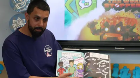 The father wearing blue is on the left of the photo holding the book in the middle of the image at the bottom. There are drawings and words in the book.