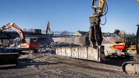 North Somerset Council A large machinery arm picks up a metal piece of a demolished bridge on a demolition site. Other construction vehicles can be seen moving rubble around the site. A temporary footbridge, replacing a gap where the bridge once stood, can be seen in the distance, and it is a sunny winter day with clear skies.