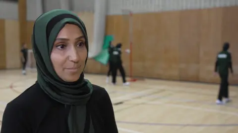 BBC Sadia Hussain wears a dark green hijab and a black top. She is standing on the left with three netball players visible in the blurred background.